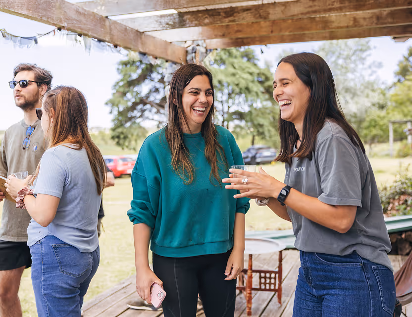 Four young adults standing outdoors under a wooden pergola, two women in front laughing and chatting while holding drinks.