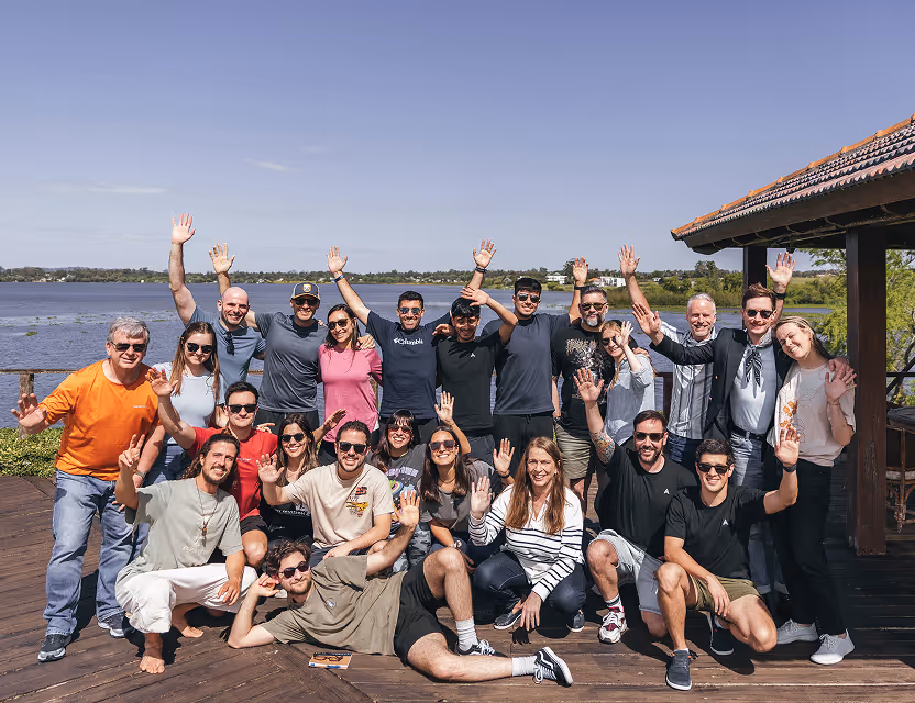 Group of diverse adults smiling and waving on a wooden deck by a lake under a clear sky.