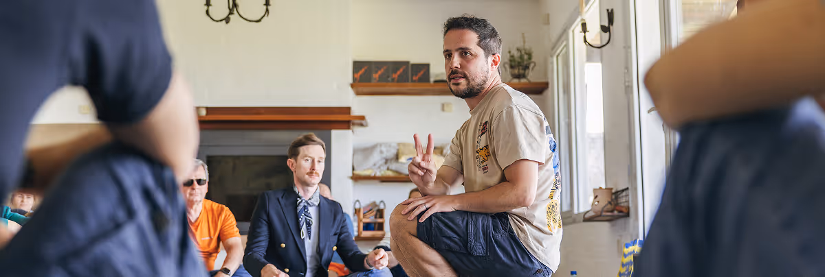 Man in casual clothes squatting and speaking to a seated group in a bright room with shelves and windows.