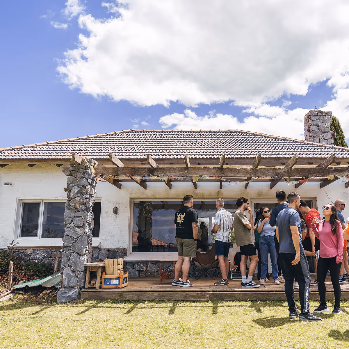 Group of people socializing on a wooden patio outside a stone house under a partly cloudy sky.