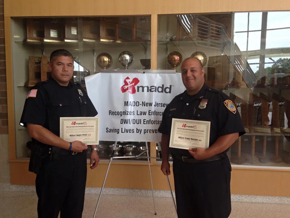 Two police officers in uniform holding certificates standing beside a MADD-New Jersey acknowledgment poster in a trophy display area.