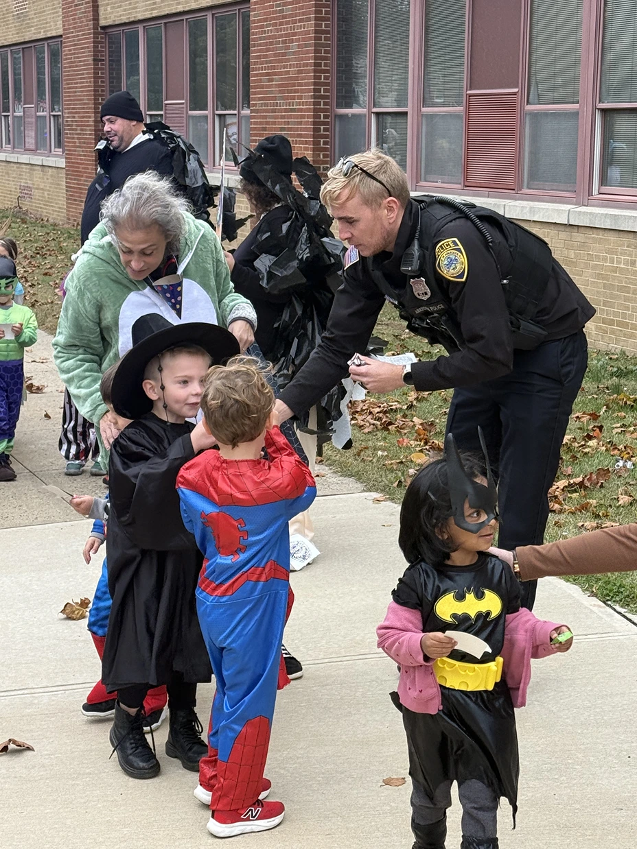 Children dressed in Halloween costumes including Spider-Man, Batgirl, and a wizard interacting with a police officer on a sidewalk.