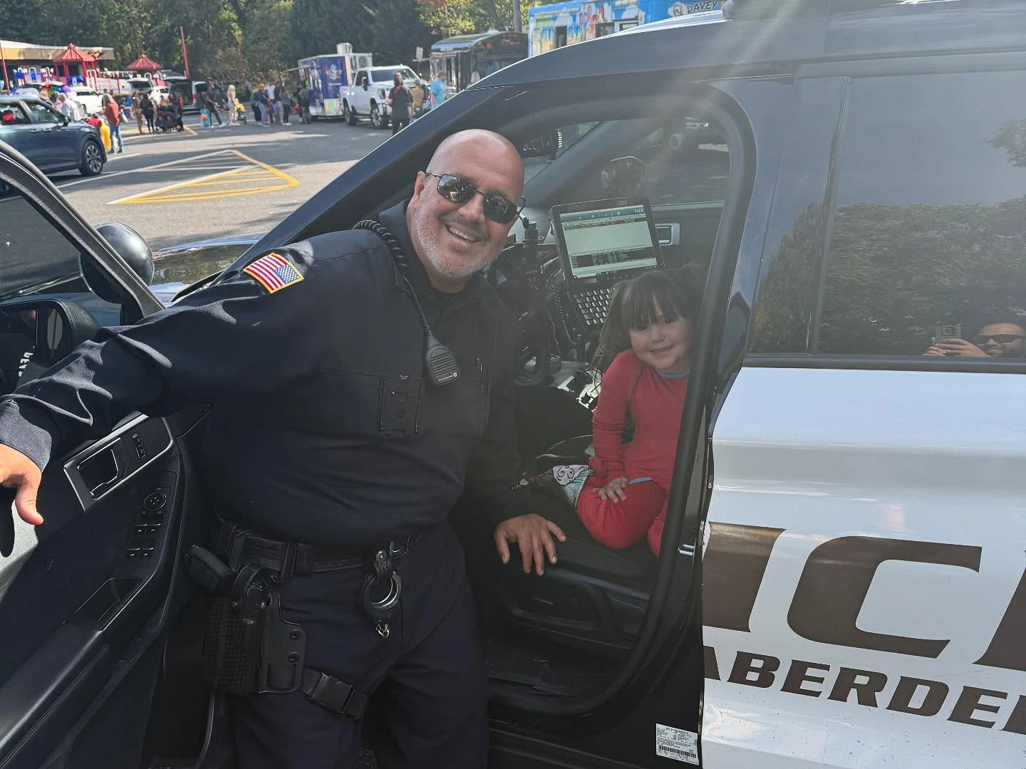 Smiling police officer standing by an open car door with a young girl sitting in the front seat of a police car.