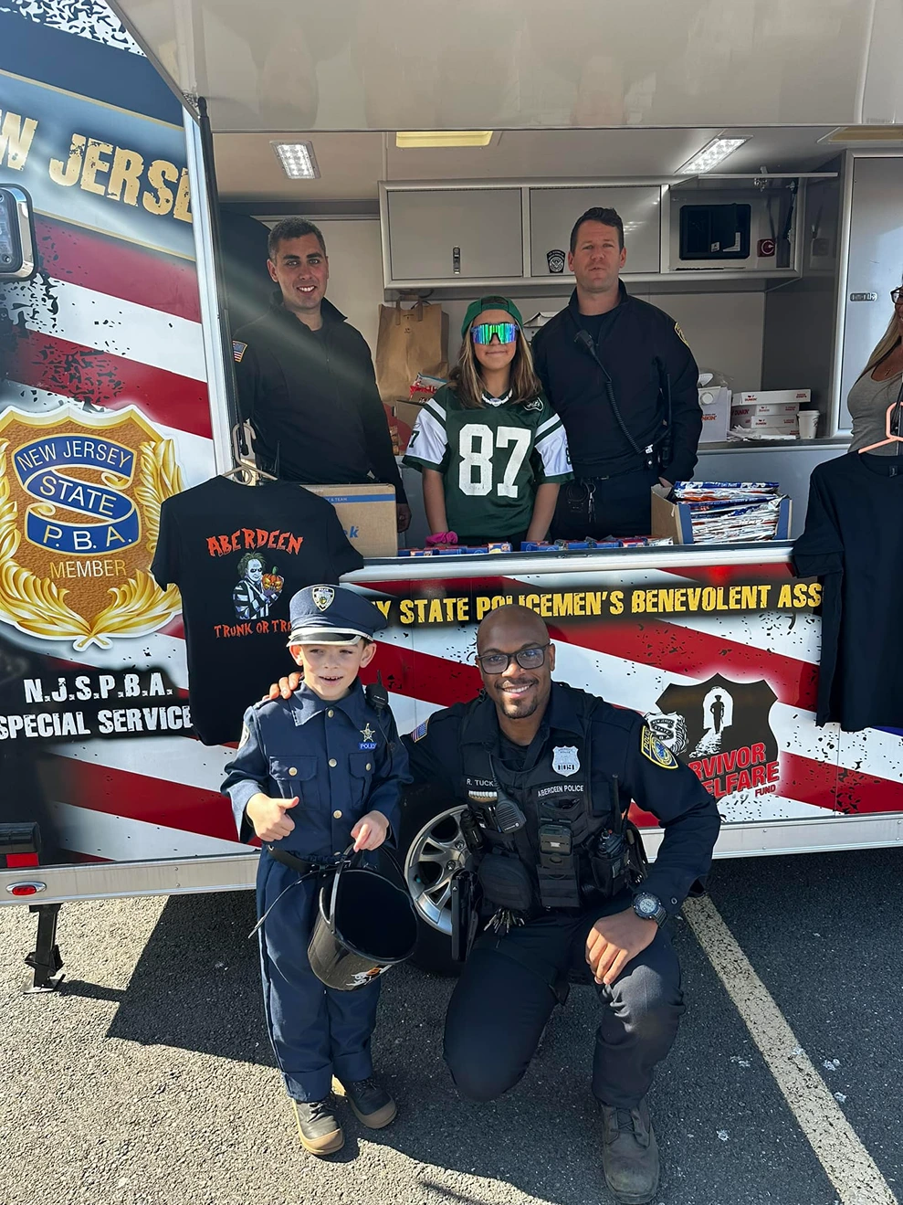 A young child dressed as a police officer stands next to a kneeling Akron police officer in front of a New Jersey State Policemen's Benevolent Association booth with two officers and a girl in a Jets jersey behind the counter.