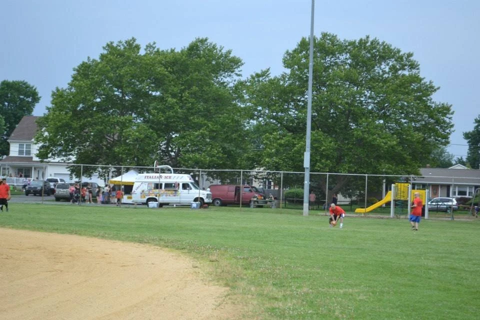 a far away view of people at the end of a baseball field and parked cars