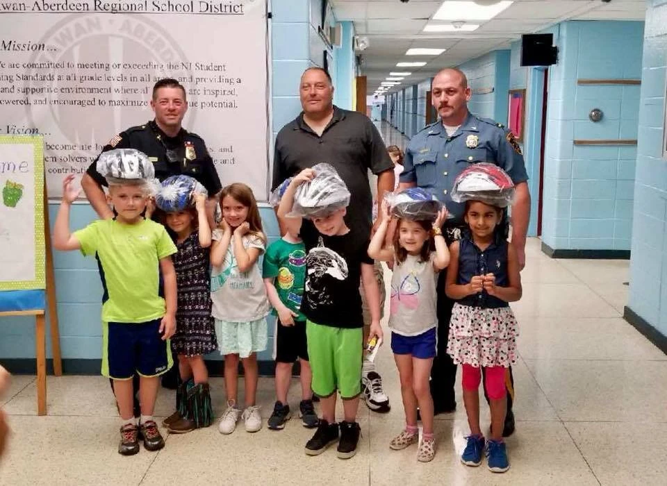 3 adults stand behind 5 children wearing bicycle helmets