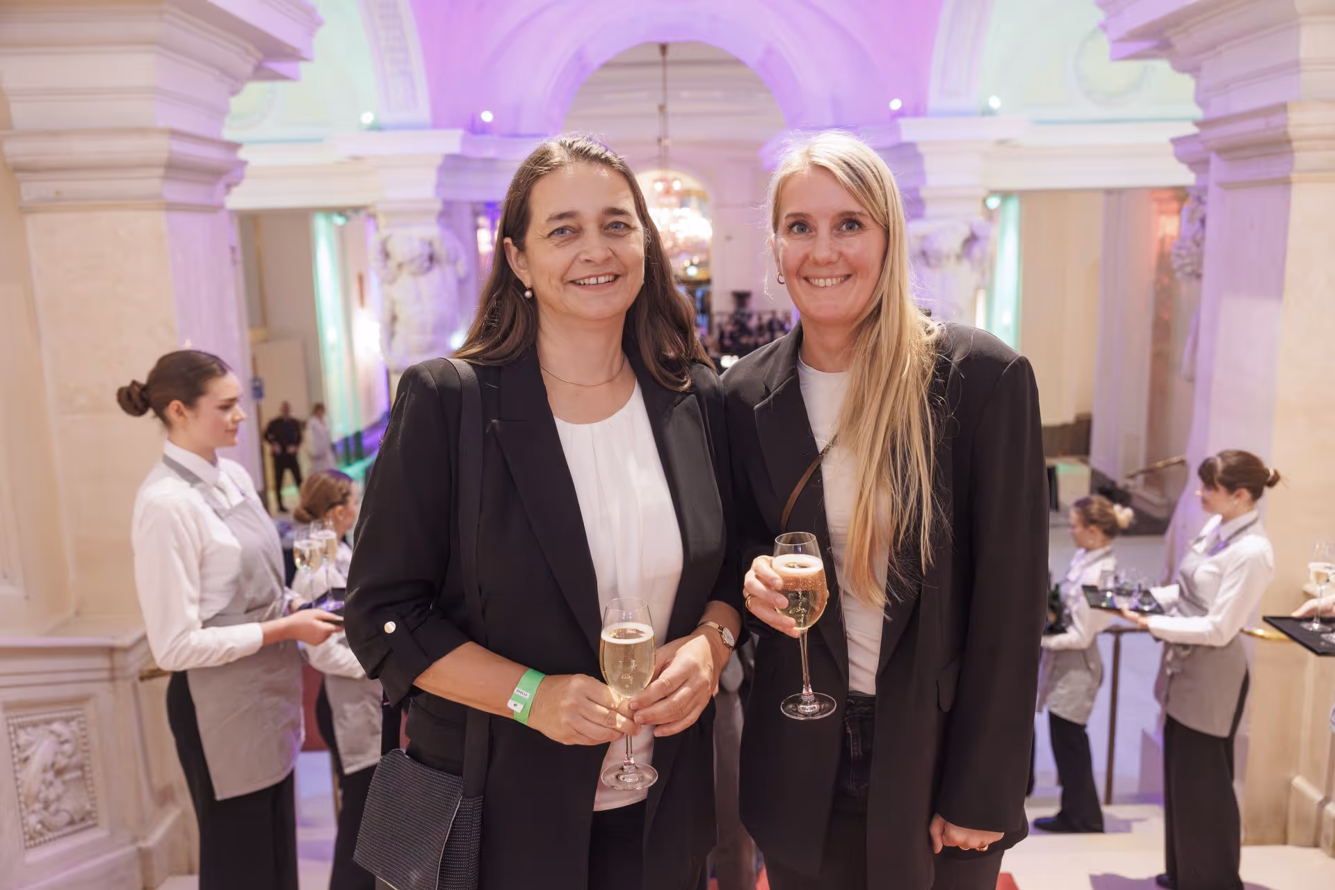 Zwei Frauen in formeller Kleidung lächeln und halten Sektgläser in einem elegant beleuchteten Veranstaltungsraum.