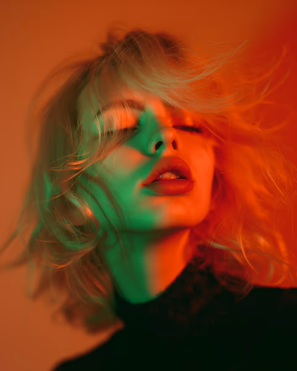 Close-up of a woman with blonde hair blowing across her face, illuminated by red and green lighting.