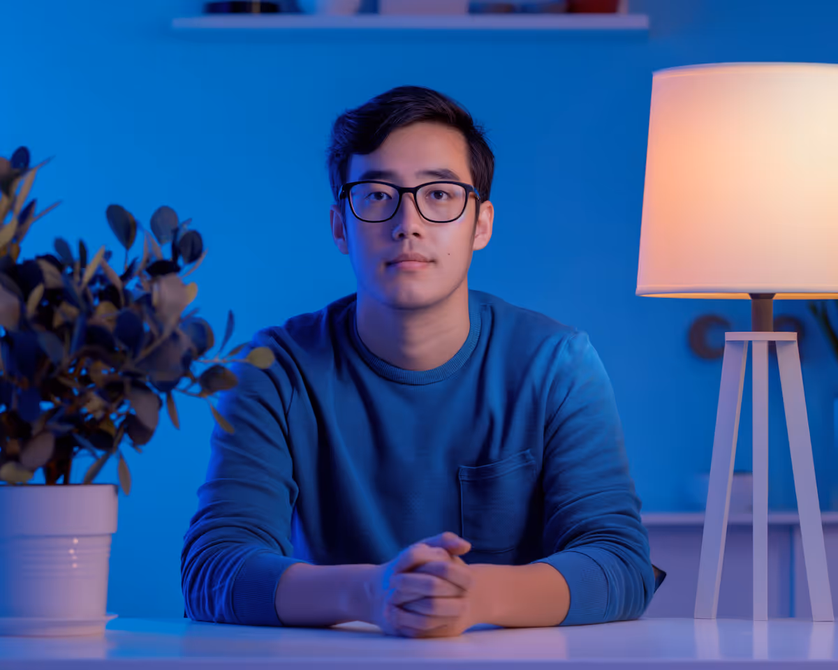 Man wearing glasses and a blue sweater sitting at a table with folded hands, next to a lit lamp and a potted plant.
