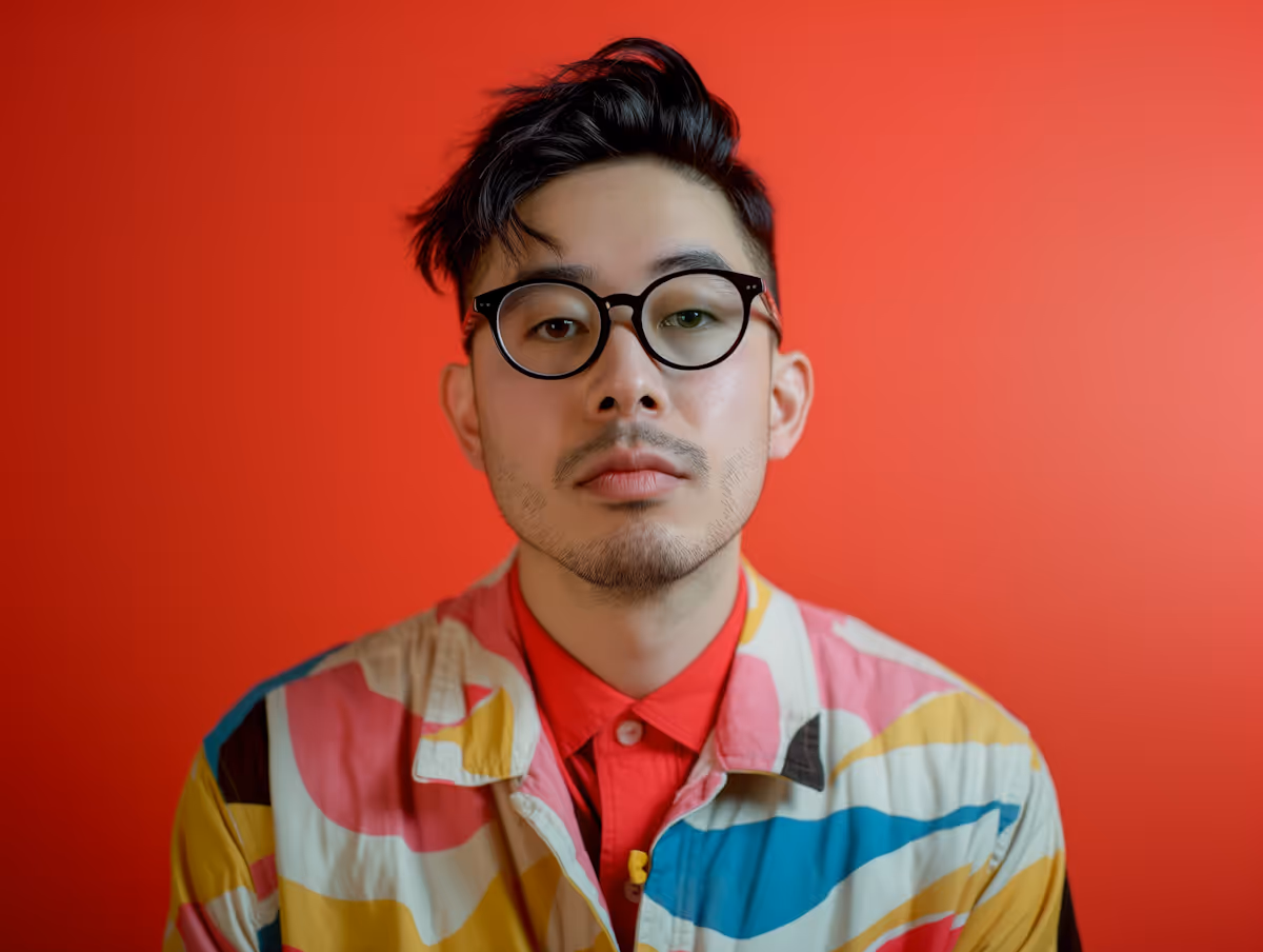 Man with black hair and glasses wearing a colorful patterned shirt and red background.