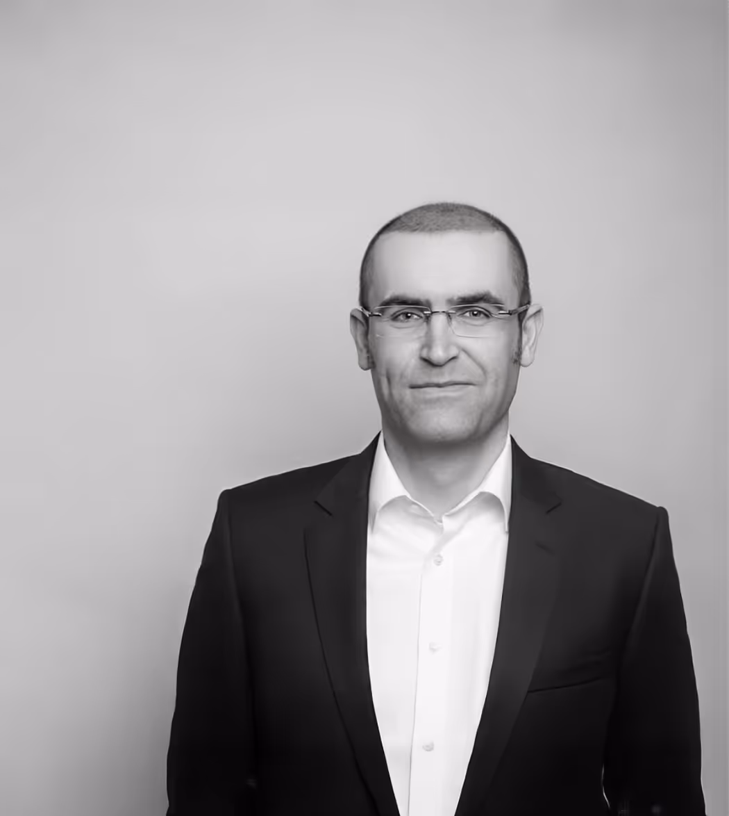 Portrait of a smiling Marius Bauer wearing glasses, a white shirt, and a dark suit jacket against a plain background.