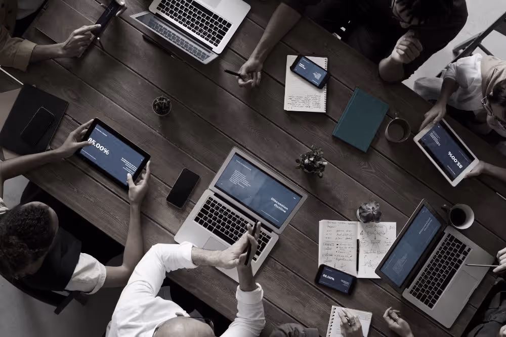Overhead view of people working at a wooden table with laptops, tablets, smartphones, notebooks, and coffee cups during a meeting.