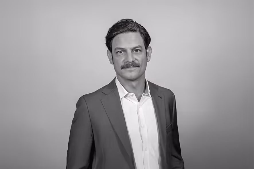 Dirk Sturz with dark hair and mustache wearing a suit jacket and white shirt against a plain background.