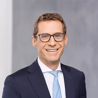 Smiling Nick Wittek with short brown hair, wearing glasses, a dark suit, white shirt, and light blue tie.