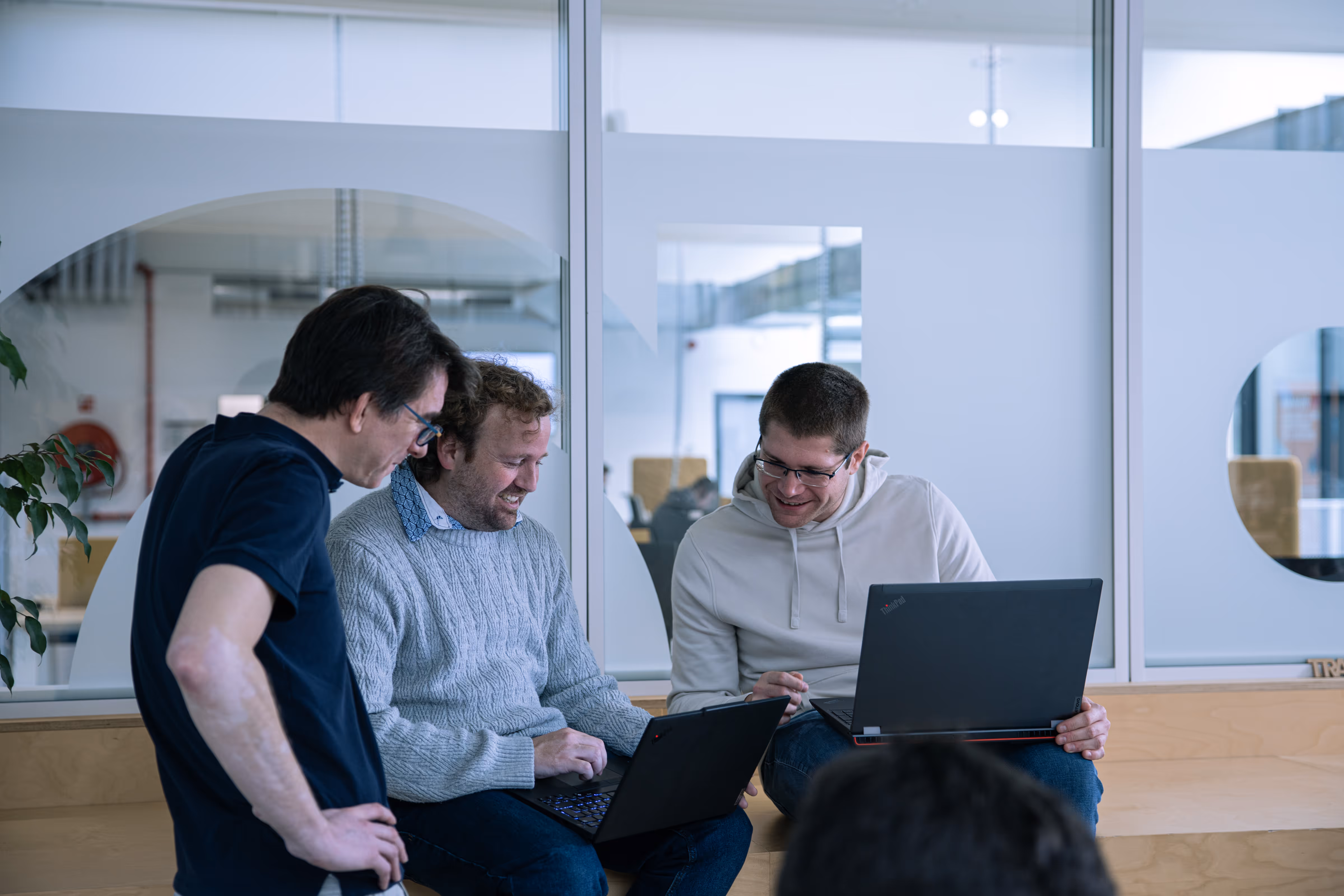 Trois hommes discutent et sourient en consultant leurs ordinateurs portables dans un espace de bureau moderne.