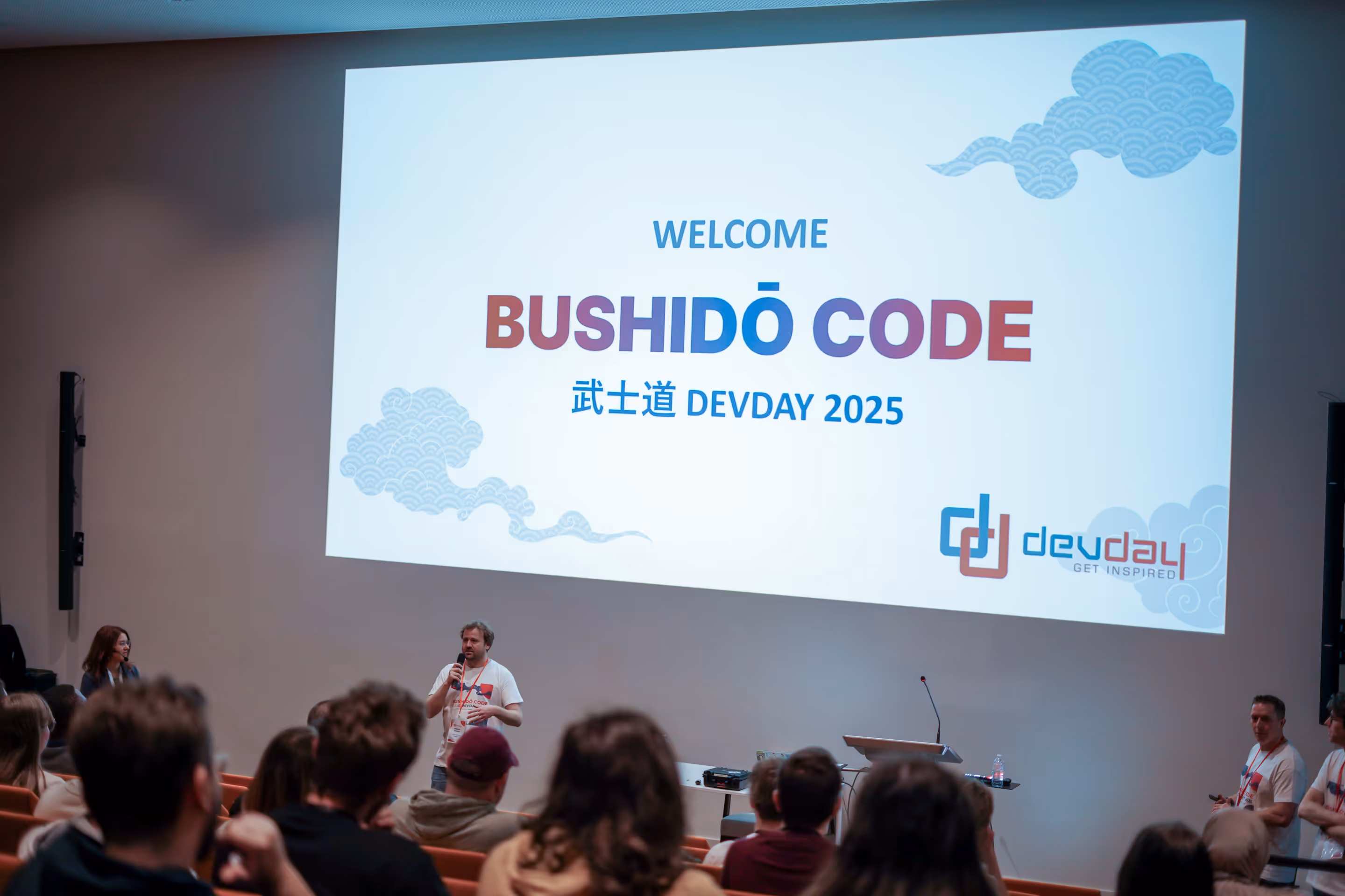 Conférencier debout devant un écran projetant le message 'Welcome Bushidō Code DEV DAY 2025' lors d'une présentation devant un public assis.