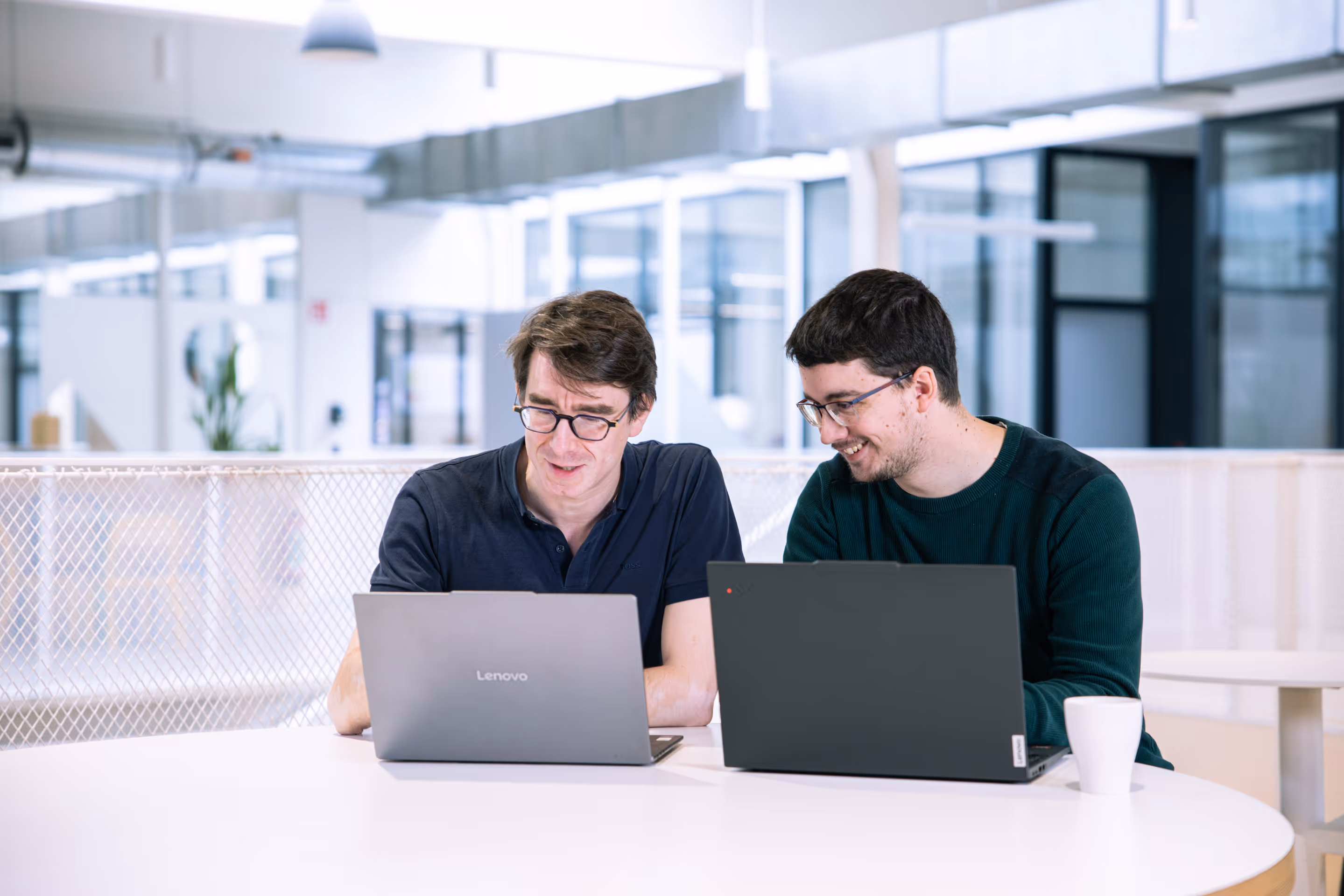 Deux hommes portant des lunettes travaillant ensemble sur des ordinateurs portables Lenovo dans un bureau moderne lumineux.