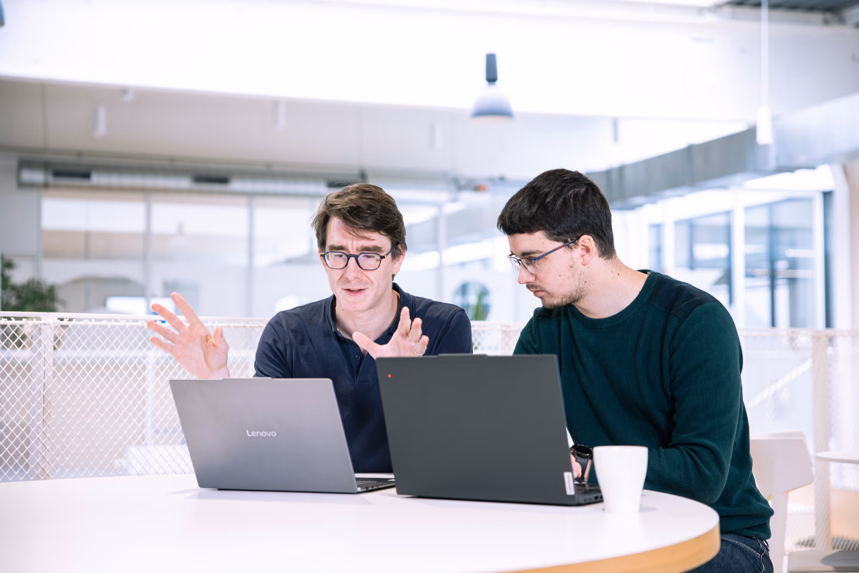 Deux hommes portant des lunettes discutent en travaillant sur des ordinateurs portables à une table blanche dans un bureau moderne.