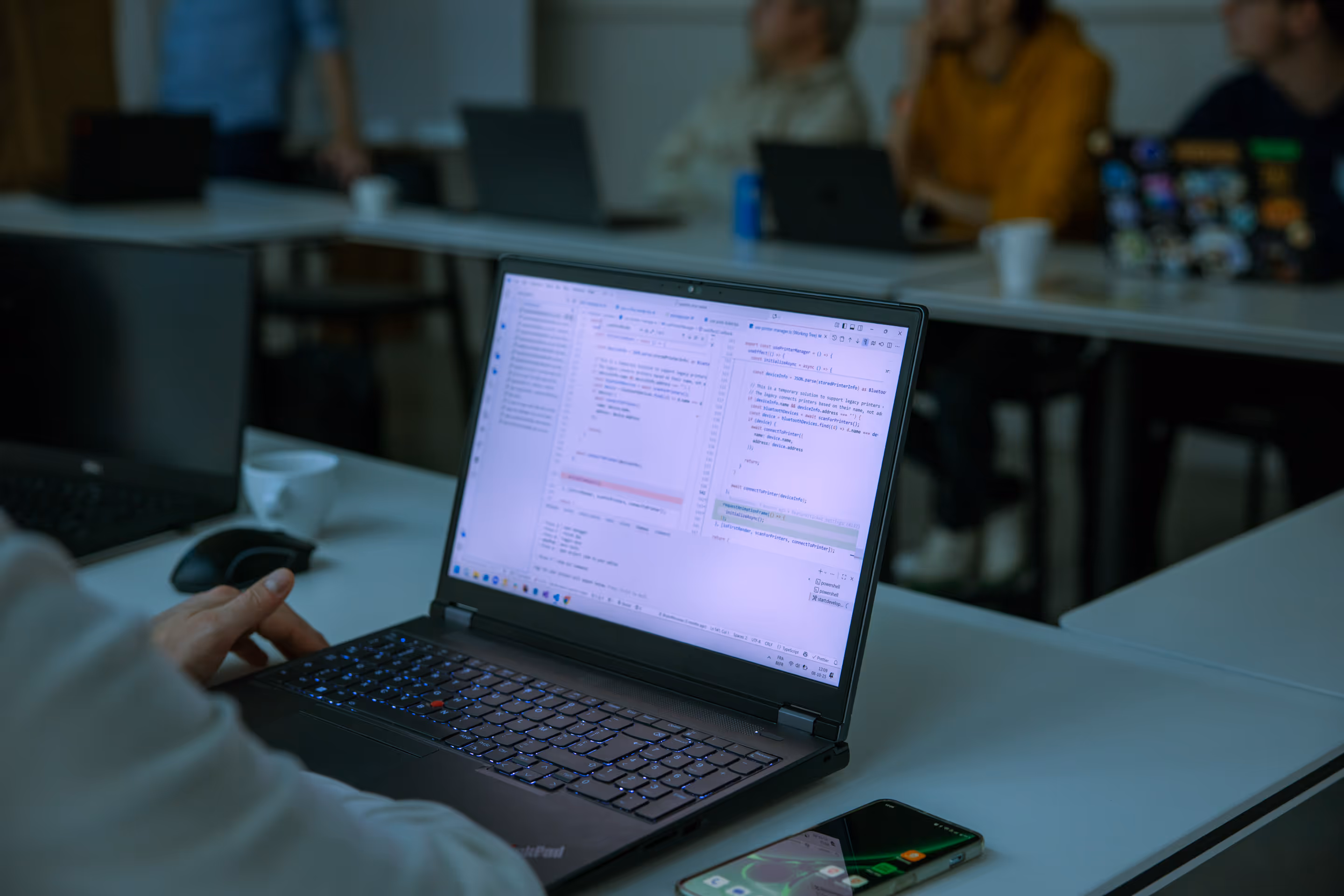 Personne travaillant sur un ordinateur portable affichant du code informatique dans une salle de réunion avec plusieurs personnes en arrière-plan.