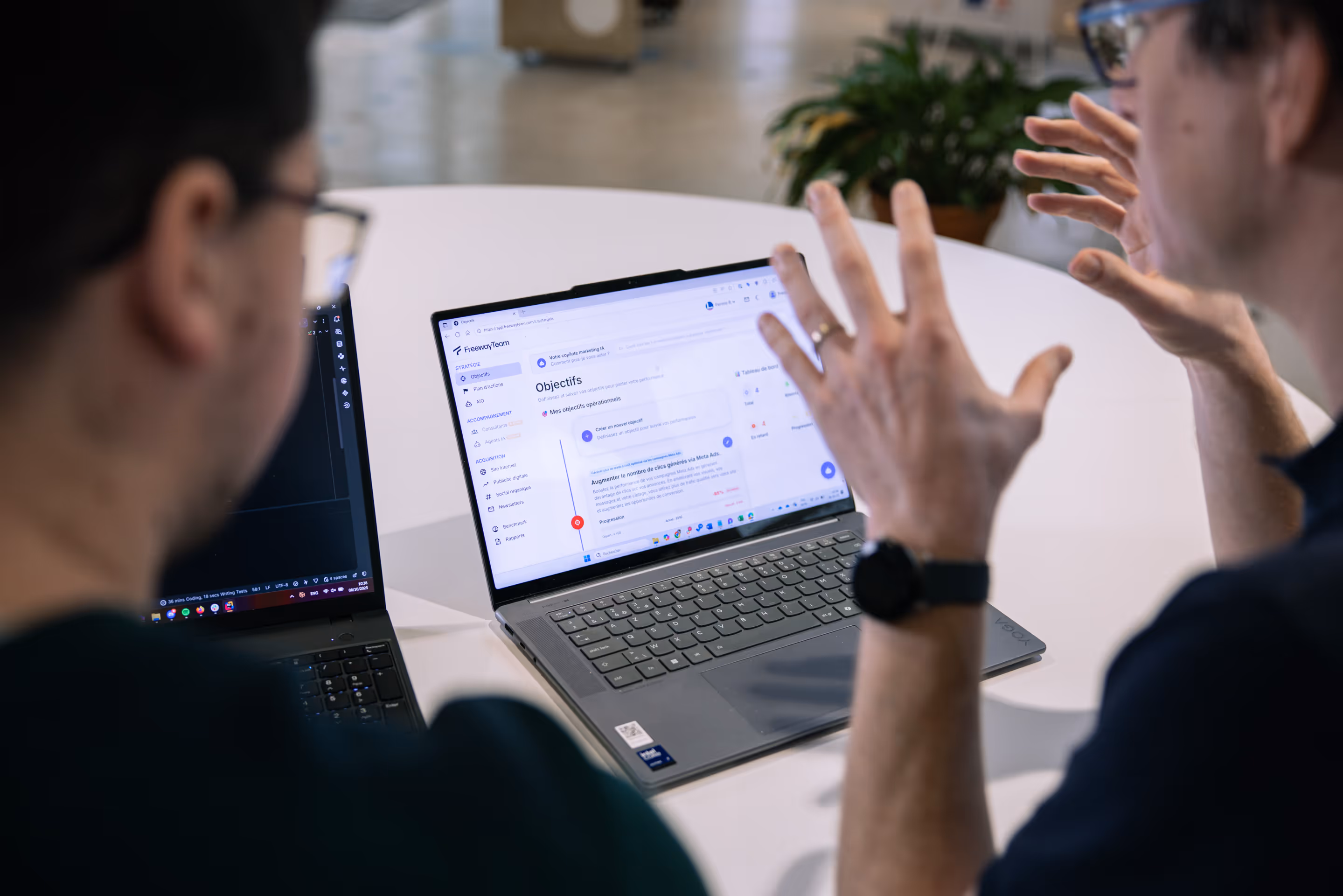 Deux personnes discutent devant un ordinateur portable affichant une interface de gestion d'objectifs marketing.