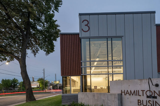 Evening view of a modern commercial building with large glass windows, illuminated interior offices, and Hamilton Central Business Park signage along a city street.