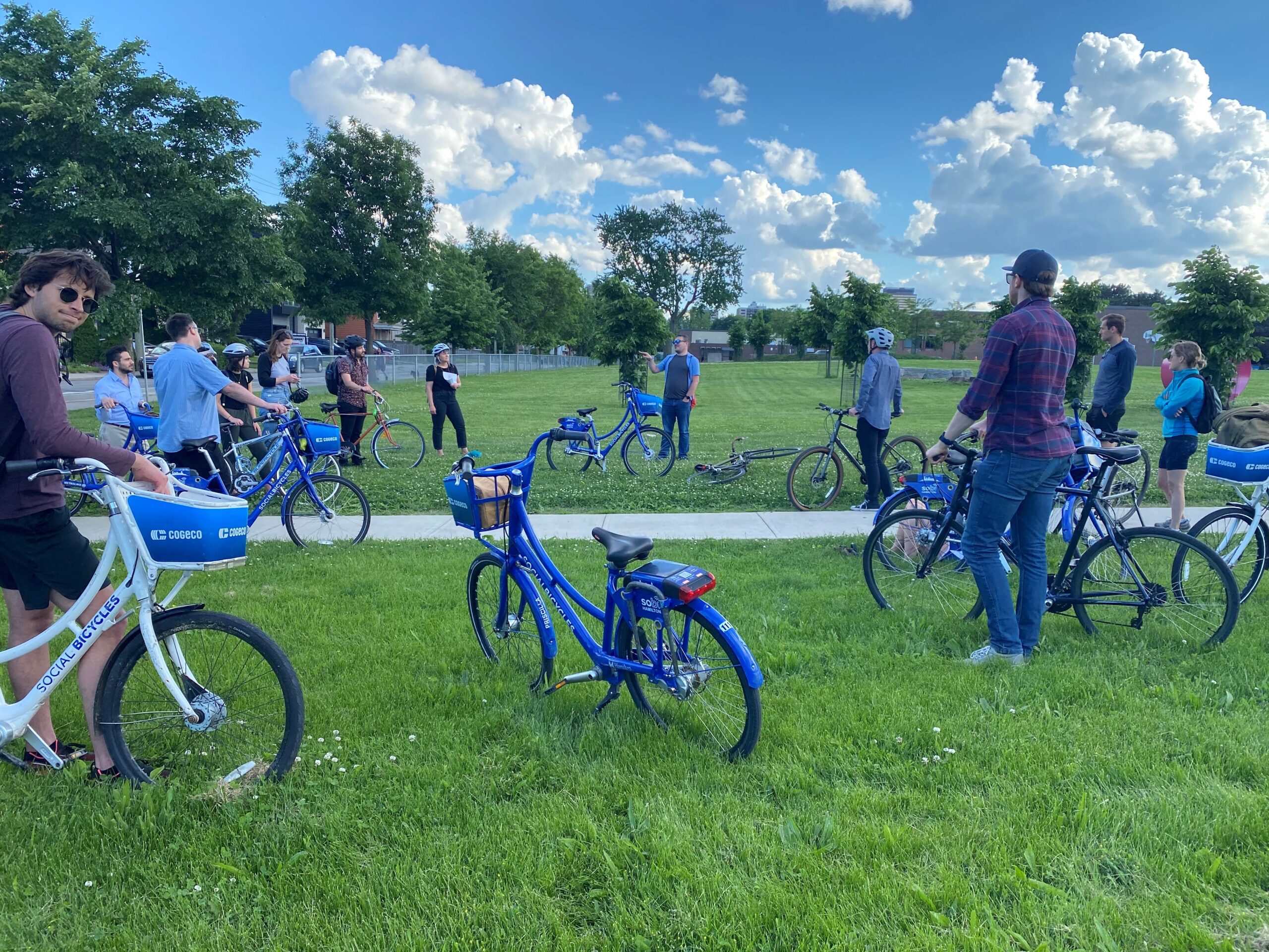 A group of cyclists gathers in a grassy park area with blue Sobi bikes, listening to a guide before starting the downtown Hamilton bike tour.