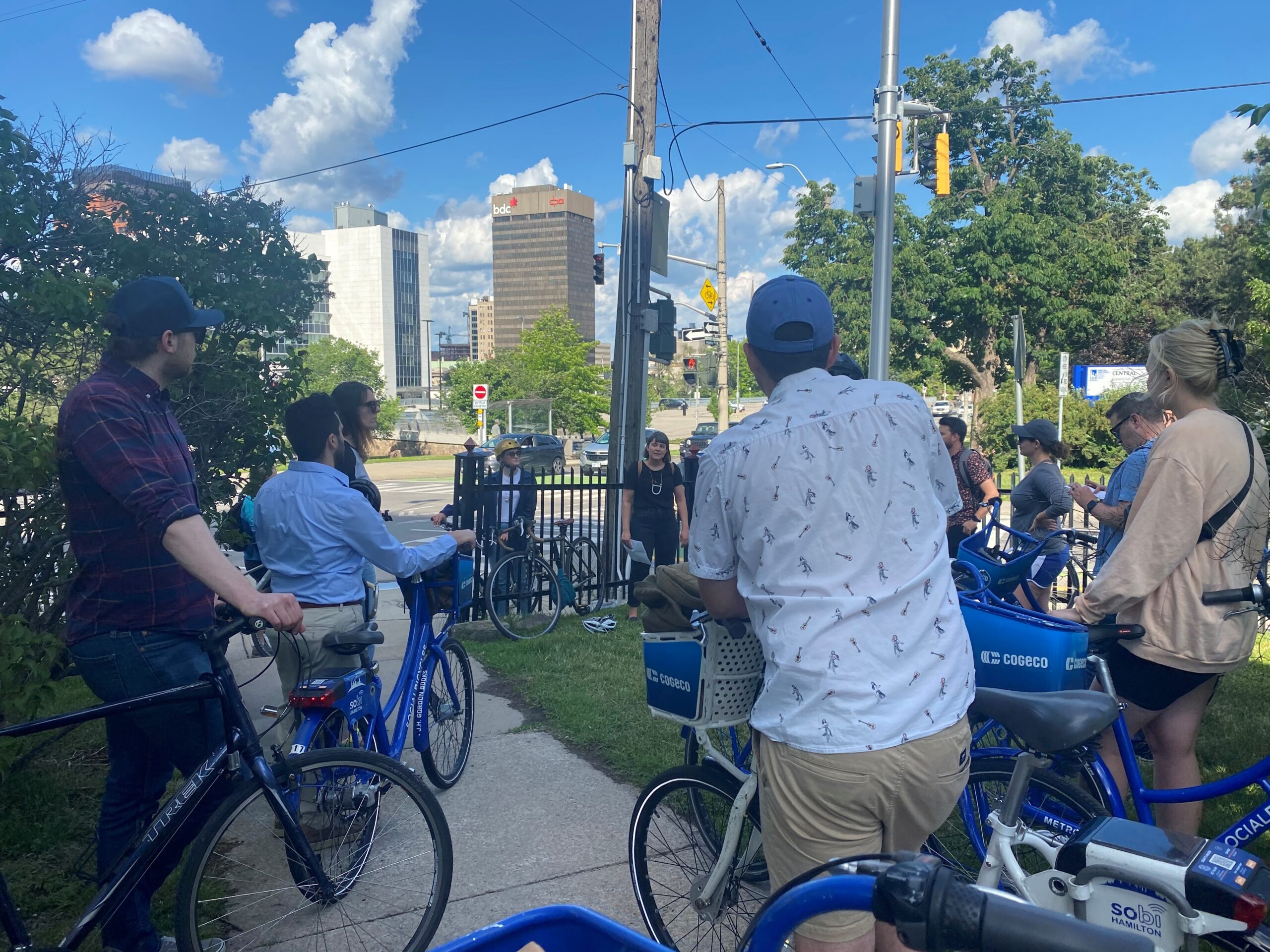 Cyclists stop at an intersection in downtown Hamilton, listening to a tour leader while city buildings and traffic signals appear in the background.