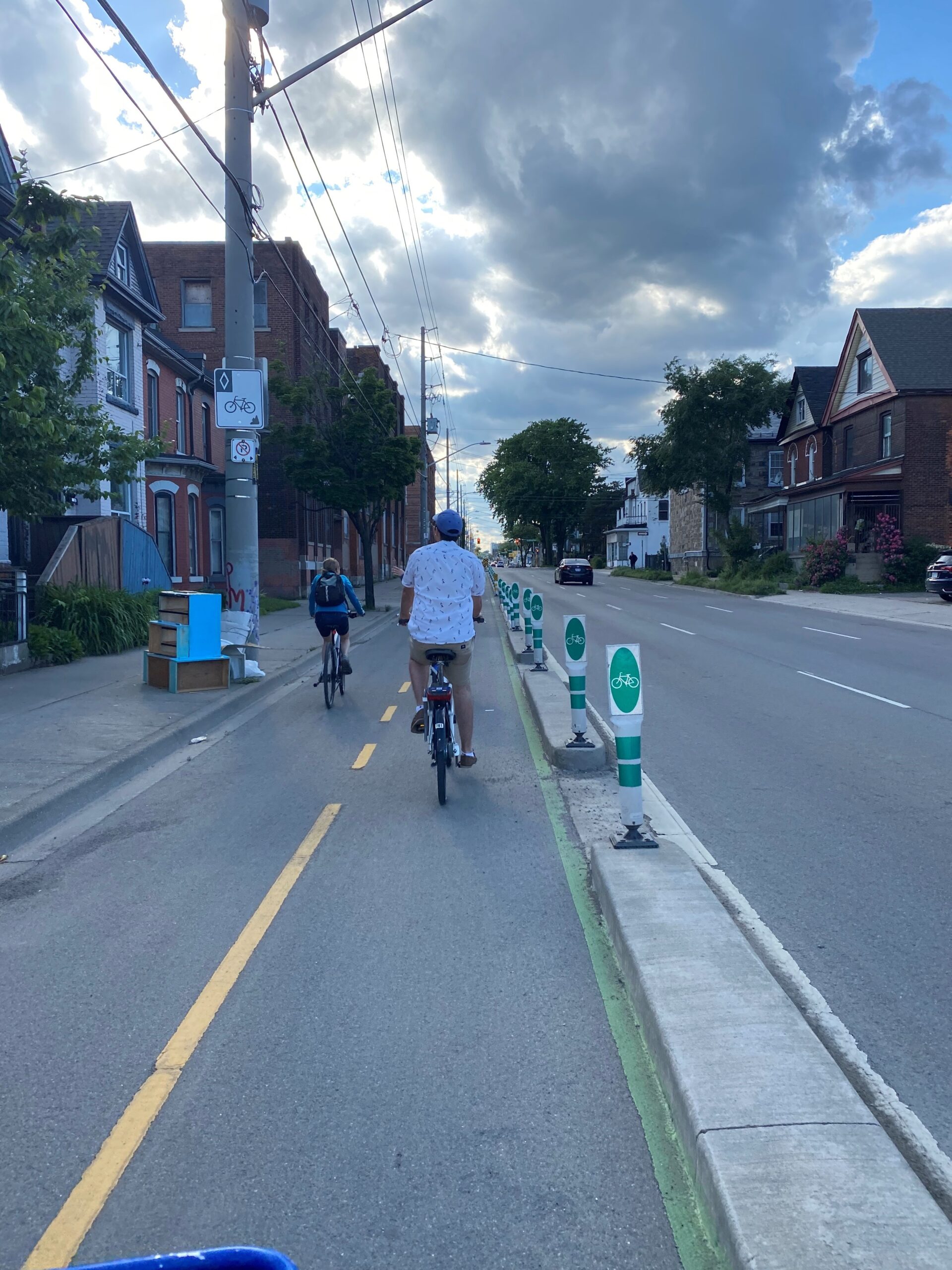 Riders travel along a protected bike lane in downtown Hamilton, passing historic homes under a cloudy sky.