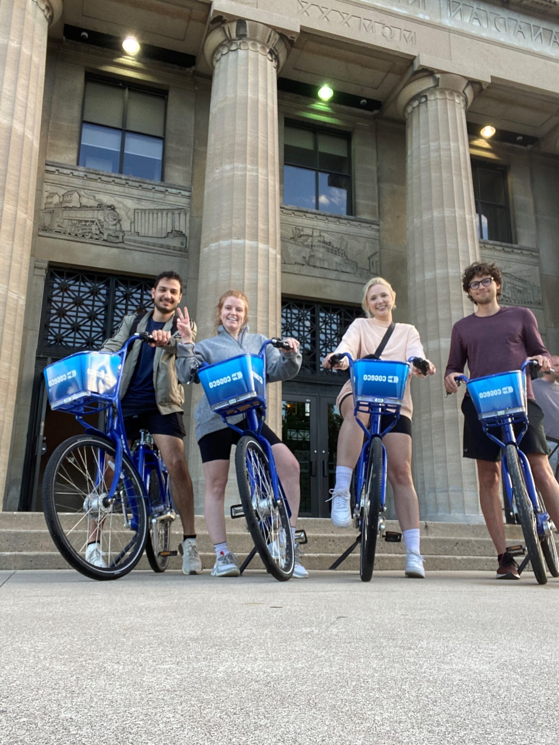 Four cyclists pose with their Sobi bikes in front of the historic LIUNA Station building in downtown Hamilton.