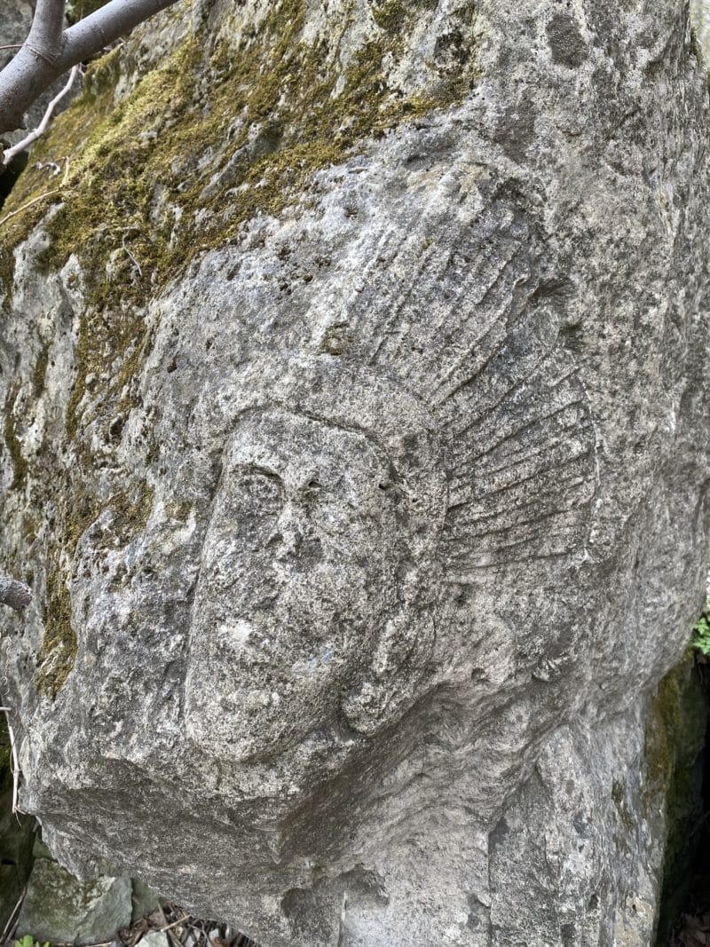 A carved face with a feathered headdress is etched into a moss-covered rock along the Cave Springs hiking trail.