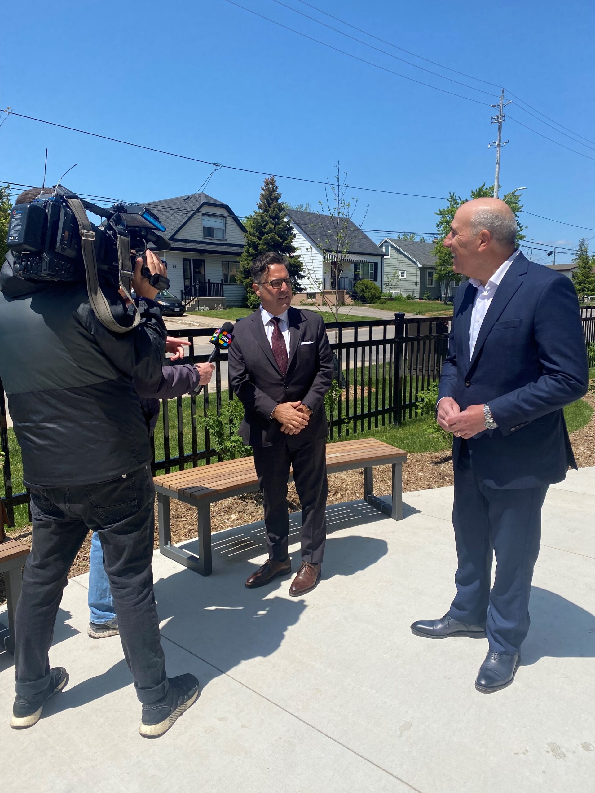 Two officials speak to a TV news camera outside 8 Roxanne Drive, standing beside newly installed benches and landscaping.