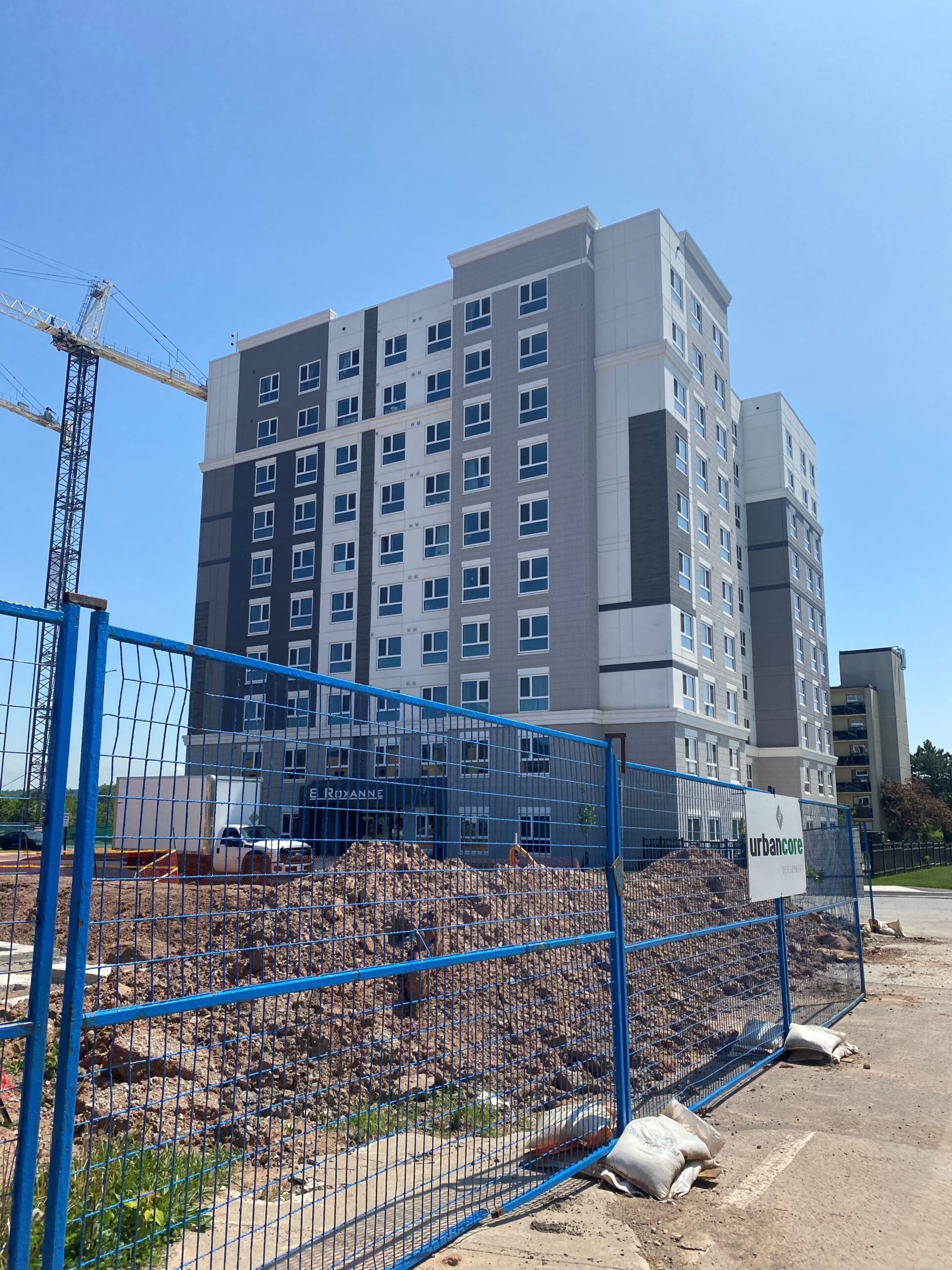 Exterior view of the completed 8 Roxanne Drive building behind construction fencing, with site work still ongoing in the foreground.