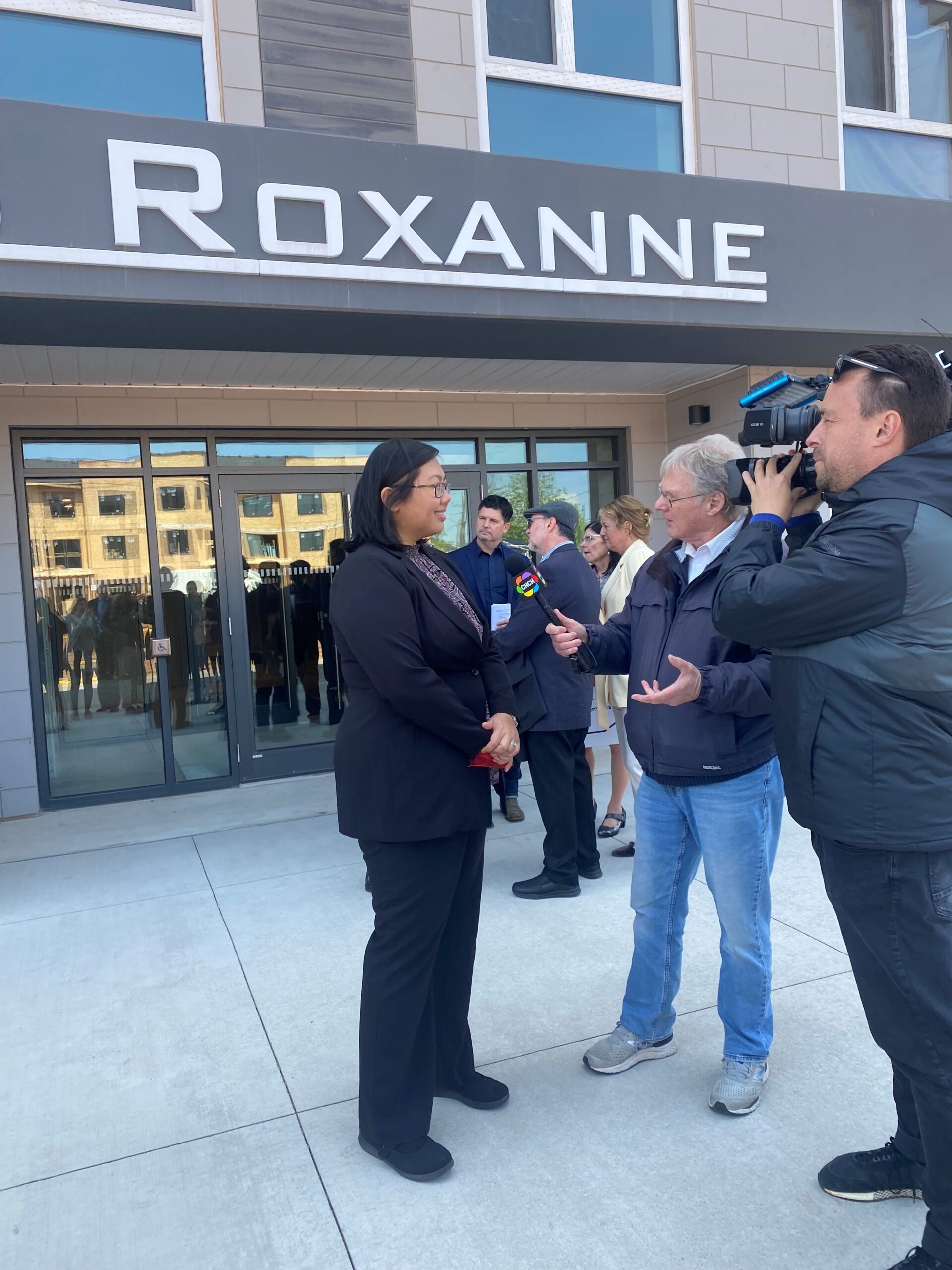 A spokesperson is interviewed by a news crew in front of the entrance to 8 Roxanne Drive, with the building’s large “Roxanne” sign visible above.