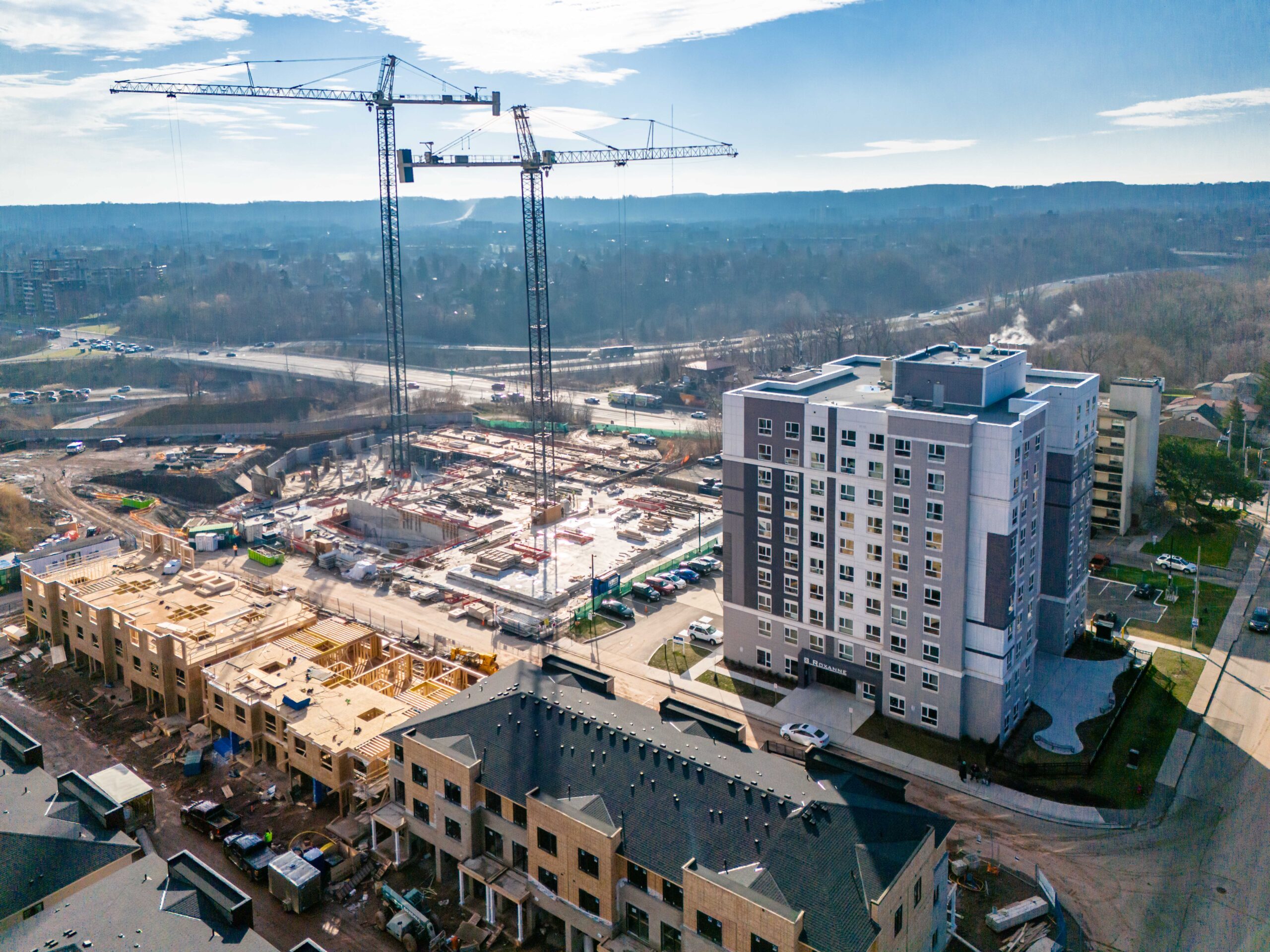 Aerial view of the Roxboro site with two tower cranes and ongoing foundation construction, surrounded by completed and in-progress residential buildings.