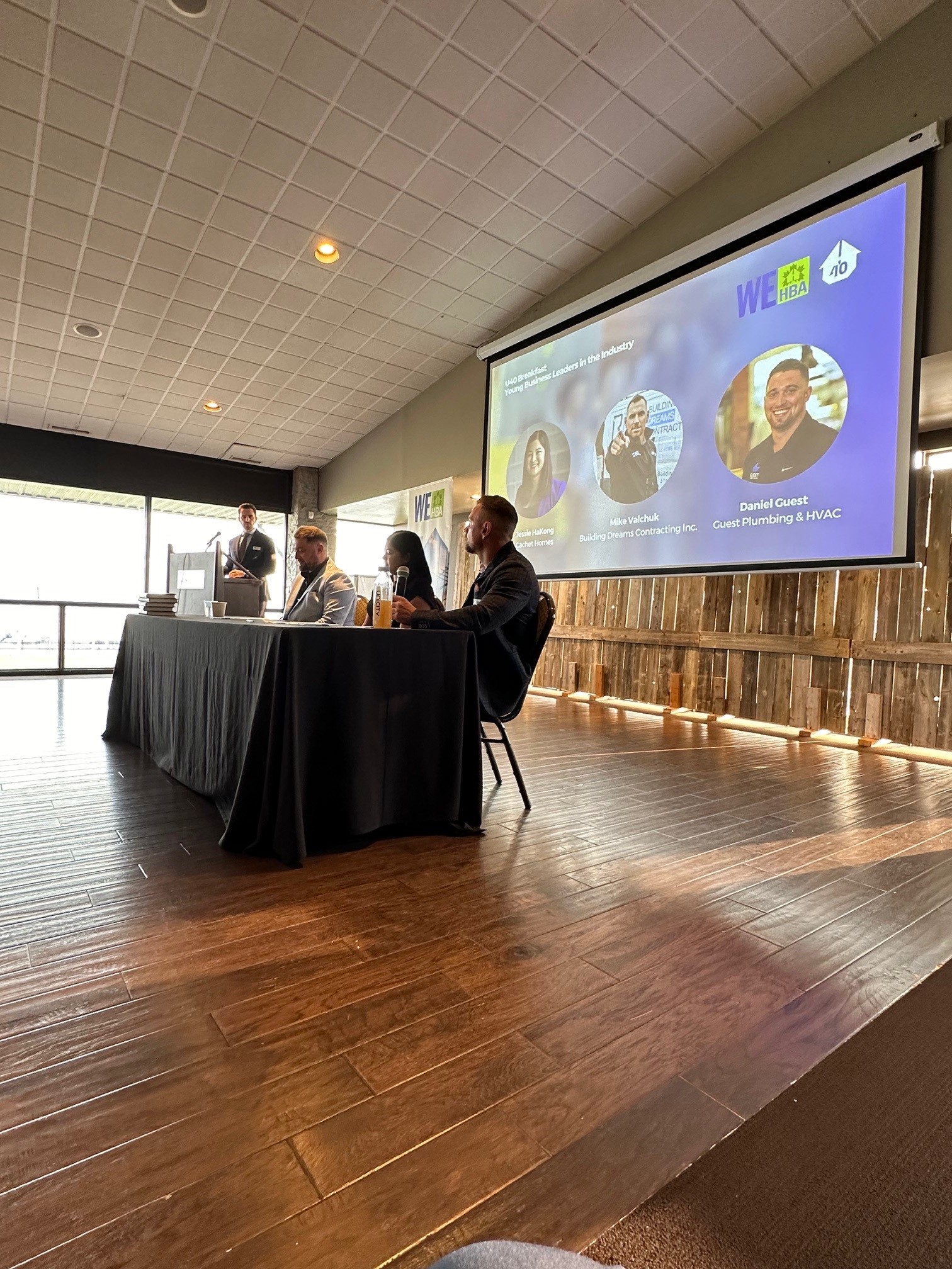 A panel of young industry leaders speaks at the WEHBA U40 Breakfast event, with a presenter at the podium and a large screen displaying the panelists’ names and photos. The audience views the discussion from a conference room with wood flooring and natural light.