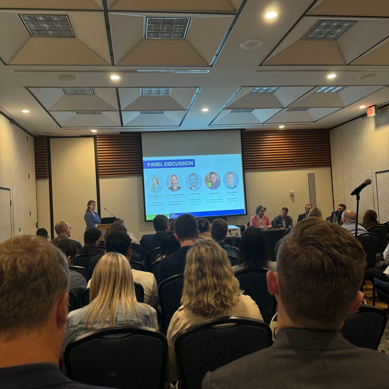 A panel discussion takes place at the OPPI Conference, with a presenter at the podium and audience members watching the panelists on stage.