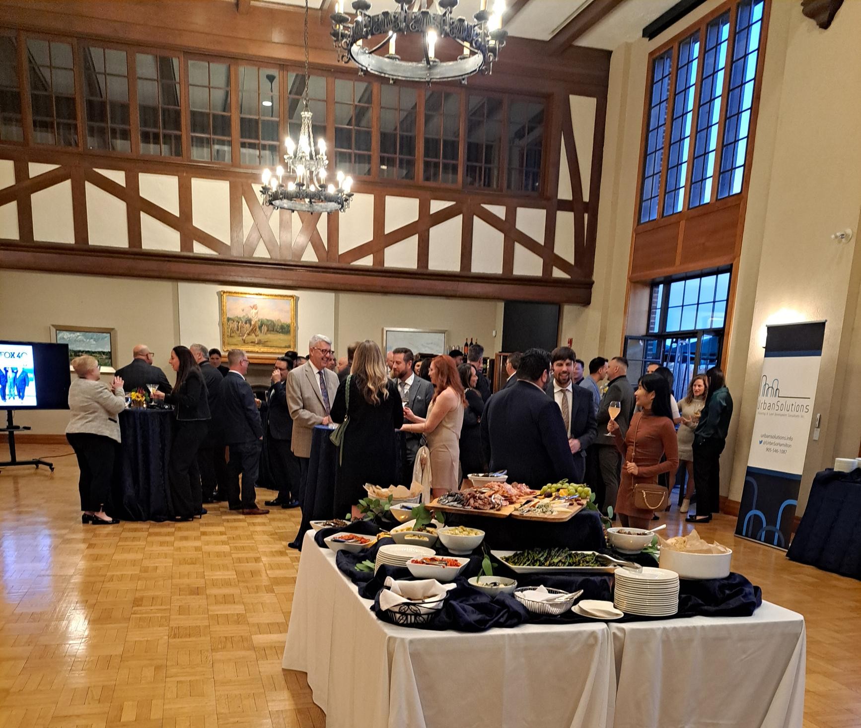 Guests gather and mingle in a large event hall during the UrbanSolutions 10-year celebration, with buffet tables and branded signage visible.