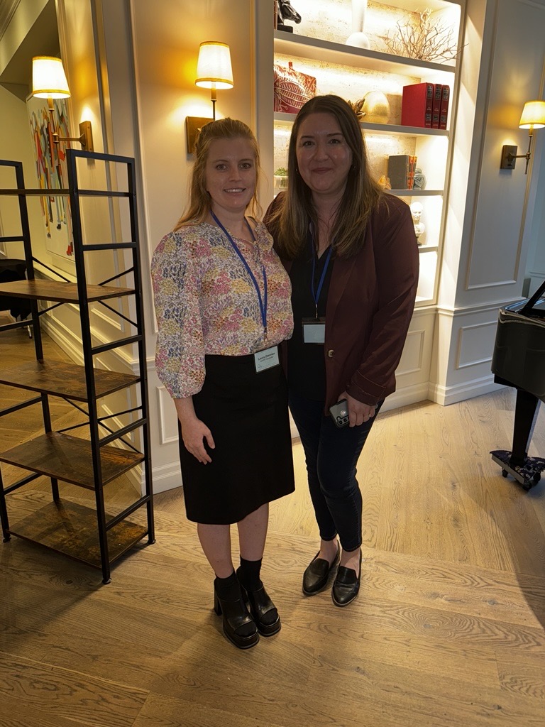 Two attendees stand together at the CACPT Conference in a warmly lit room with shelves behind them. They are both wearing conference badges and smiling toward the camera.