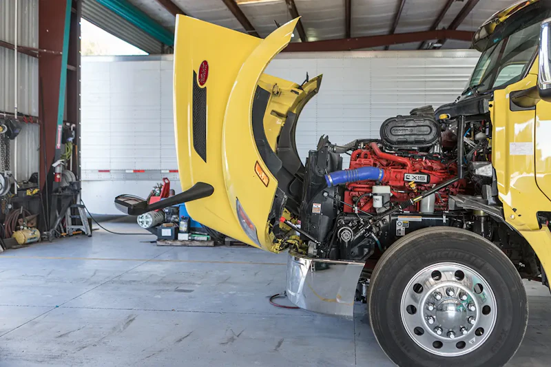 Yellow semi-truck with its hood open inside a maintenance garage, exposing a red diesel engine while tools and equipment sit in the background.