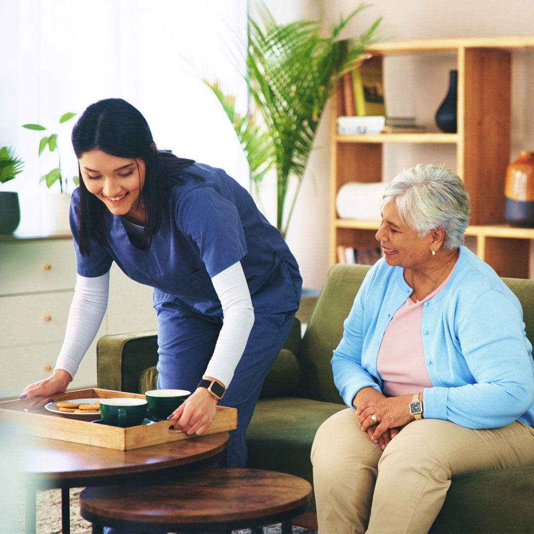Caregiver in blue scrubs smiling while placing a tray with coffee and cookies on a table for an elderly woman sitting on a couch.