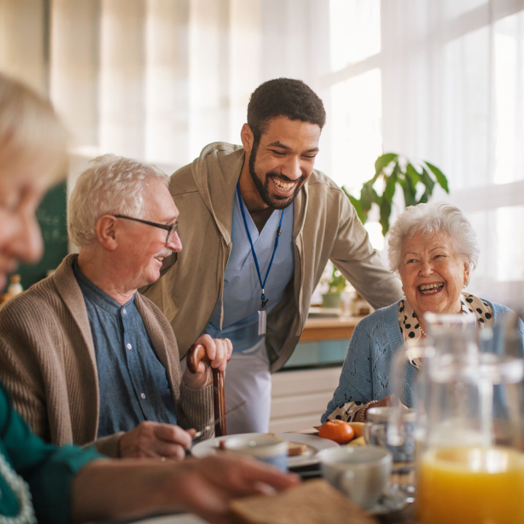 Smiling caregiver interacting with two elderly people sitting at a table with breakfast items.