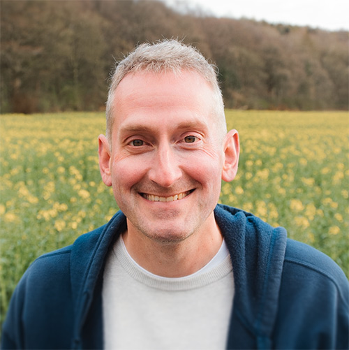 Smiling man with short gray hair wearing a blue hoodie and white shirt standing in a yellow flower field with trees in the background.