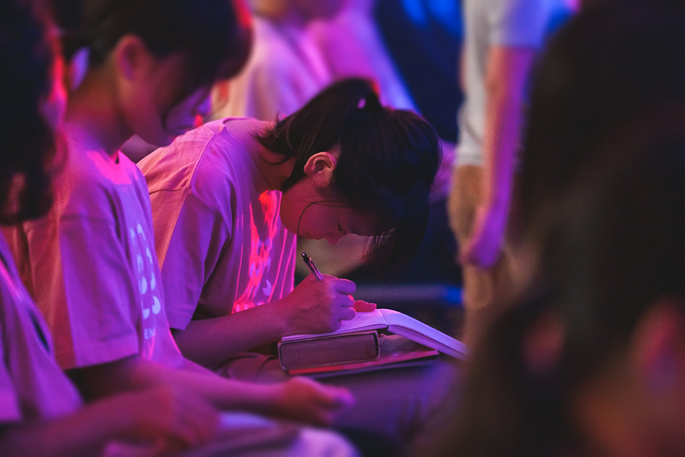 Young woman writing in a notebook during a conference with colorful stage lighting.