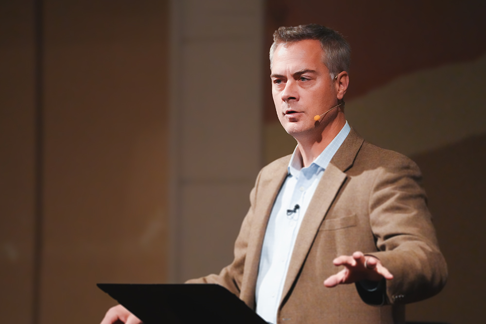 Man wearing a brown blazer and white shirt speaking with a headset microphone on stage.