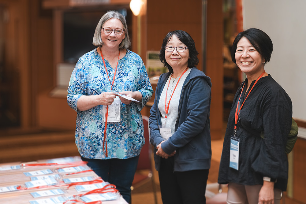 Three smiling women wearing conference badges and standing near a table with name tags at an indoor event.