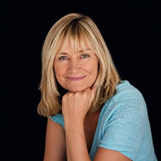 Smiling middle-aged blonde woman in a blue shirt resting her chin on her hand against a black background.