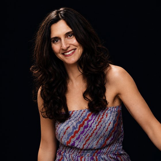 Smiling woman with long curly dark hair wearing a strapless patterned dress against a black background.