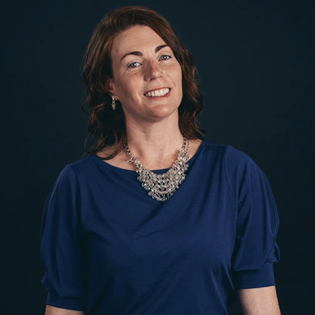 Smiling woman with medium-length brown hair wearing a blue top and a layered silver necklace against a dark background.