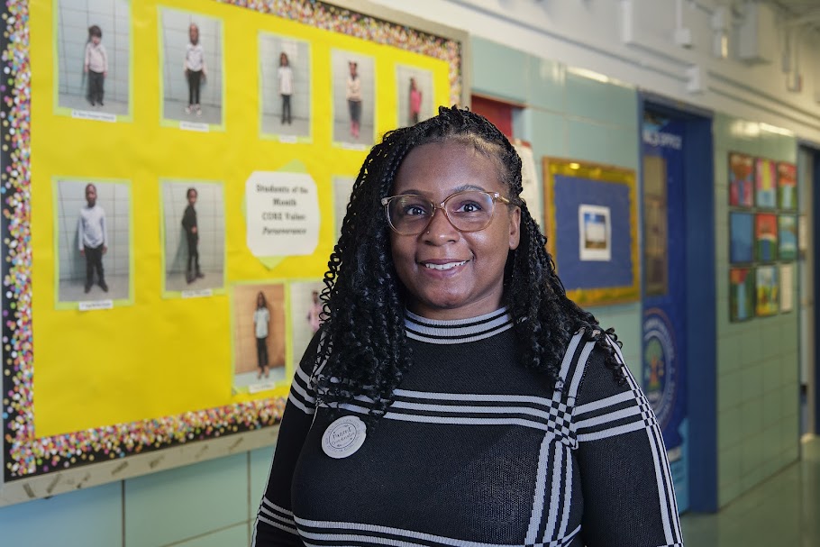 Ms. Brimage smiling in school hallway near Students of the Month bulletin board