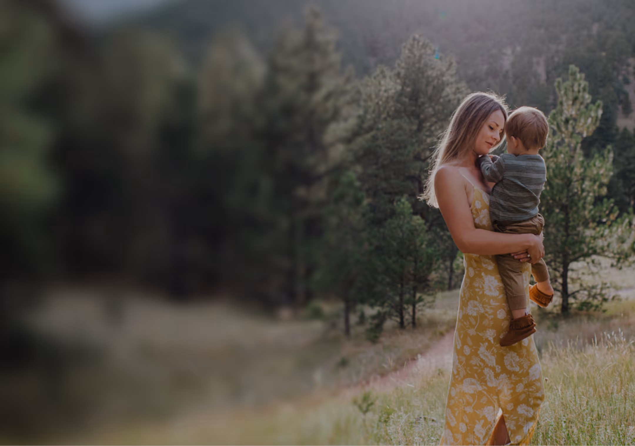 Woman in yellow floral dress holding a toddler in a grassy clearing with trees in the background.